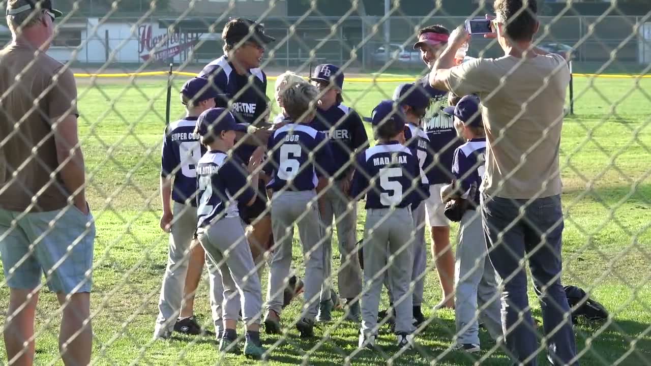 Parents watch from behind a chain-link fence as a youth baseball team huddles on the field.