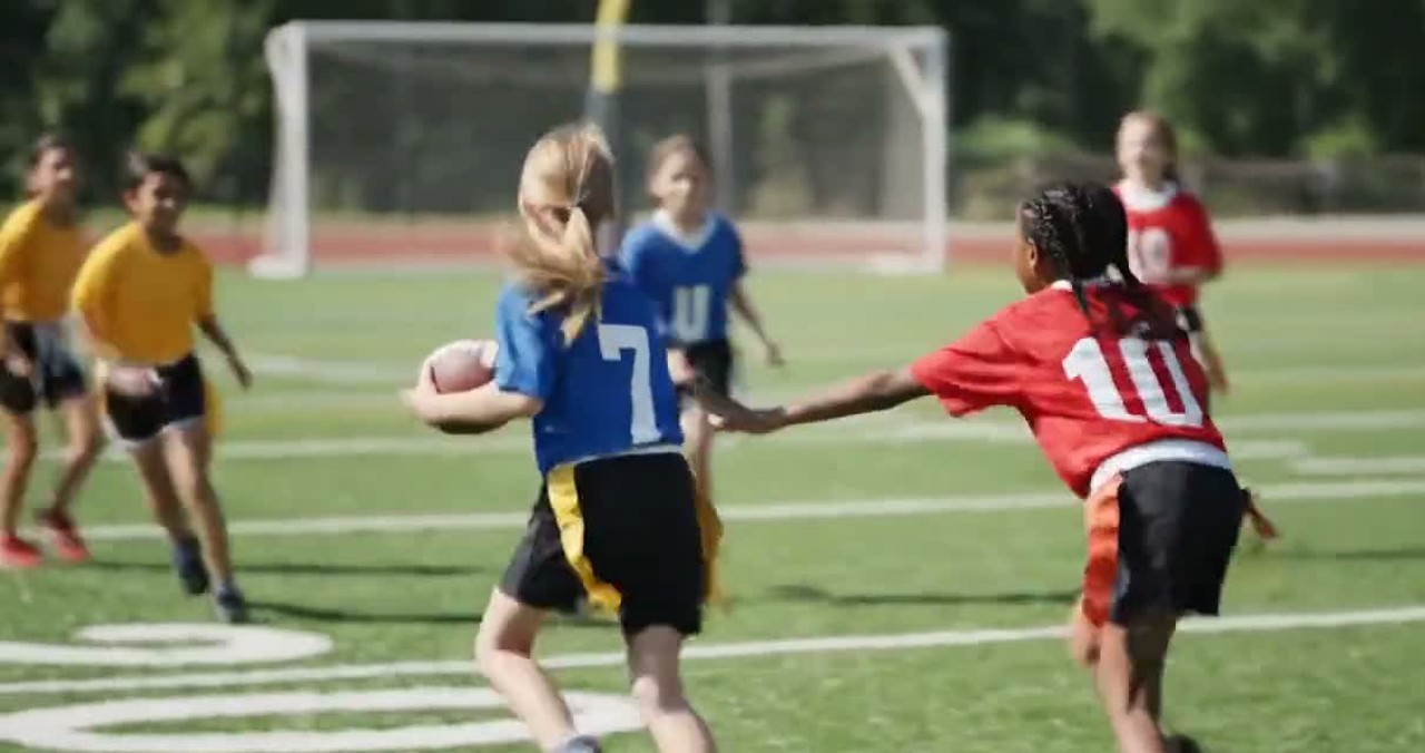 A young player carries the ball in a flag football game as a defender reaches for her flag.