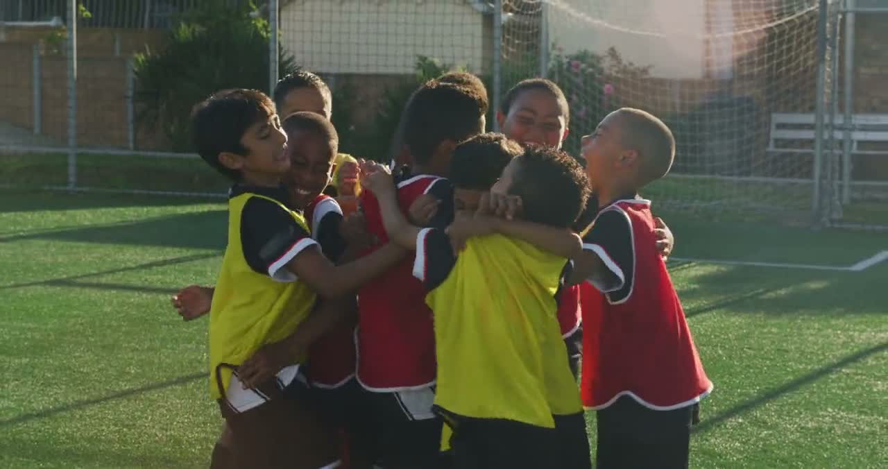 Young soccer players in yellow and red bibs hug in a group celebration on a turf field.